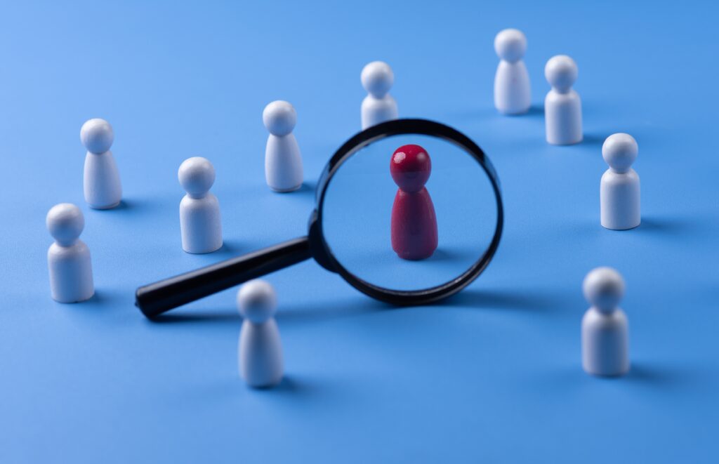 Image of peg people who are white with one that is red and highlighted with a magnifying glass.