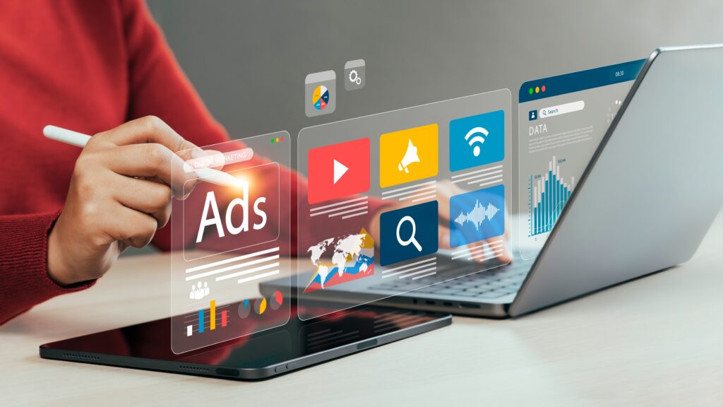 This is an image of a person sitting at a desk. There is an ipad and computer on the desk and am image is being projected from the two with the word ADS on part of the image.