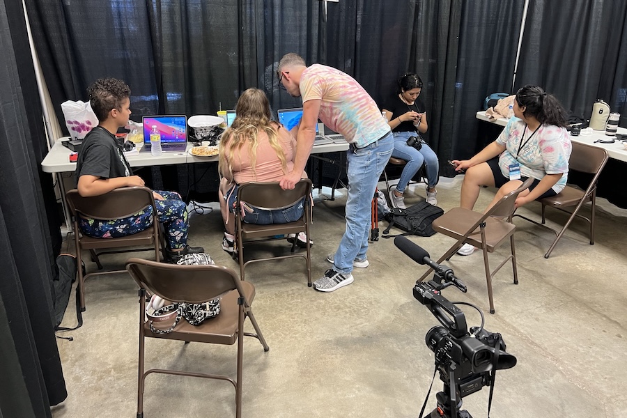 An instructor leaning over a table helping a student on a laptop. Three other students are sitting in chairs in the room.