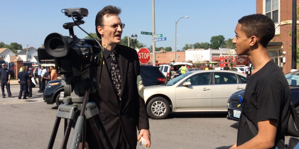 Photo of a man in a suit working a camera while talking to a kid