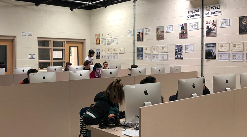 Photo of a newsroom with cubicle style desks with students at each desk. A wall shows a digital cinema all of fame showing milestones