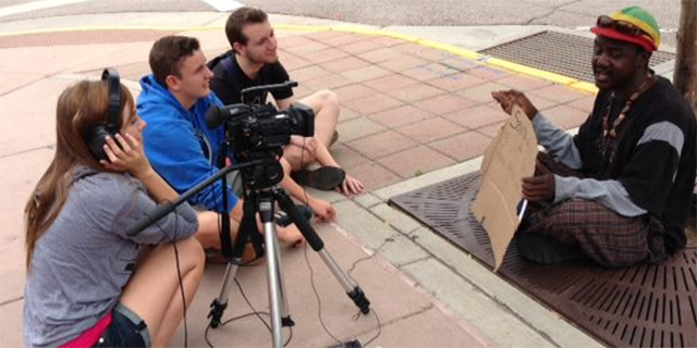 Photo of 3 people recording an interview with a homeless person sitting on the street