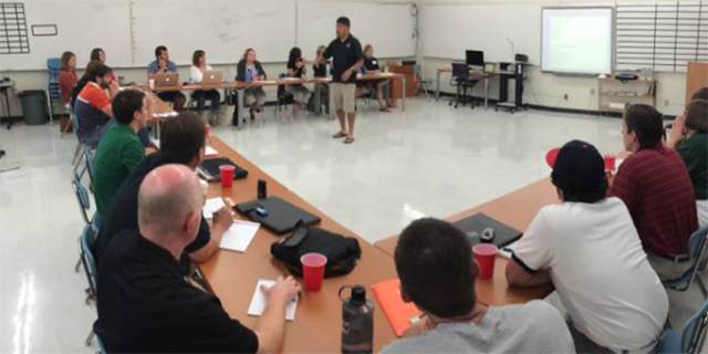 Photo of a teacher standing in the middle of desks all facing him with other teachers learning
