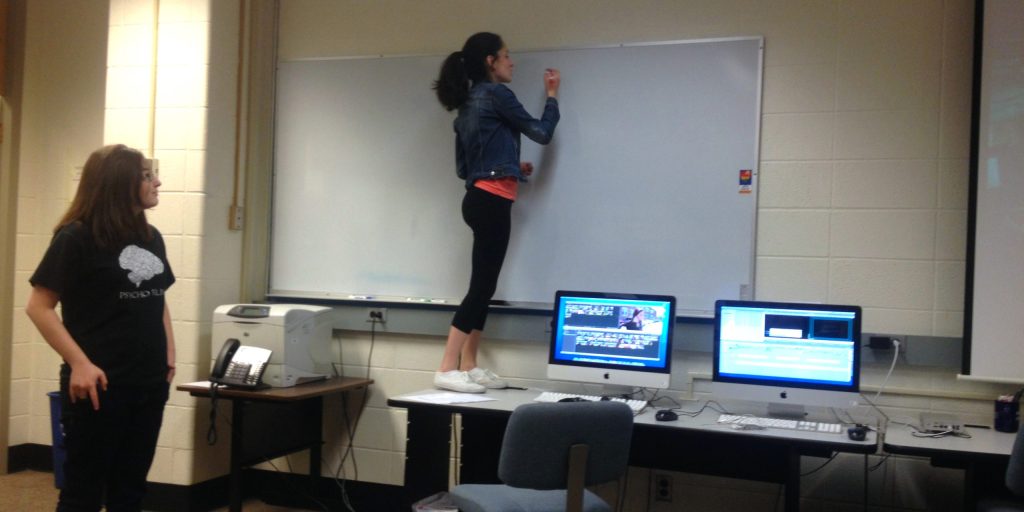 Photo of a girl standing on a desk to write on a whiteboard