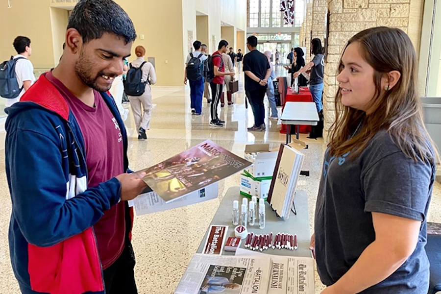 A student holds a student-made college newspaper at a booth while a girl tells him about the paper