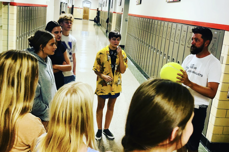 Photo of a teacher holding a giant softball and talking to 6 students in the hallway by lockers