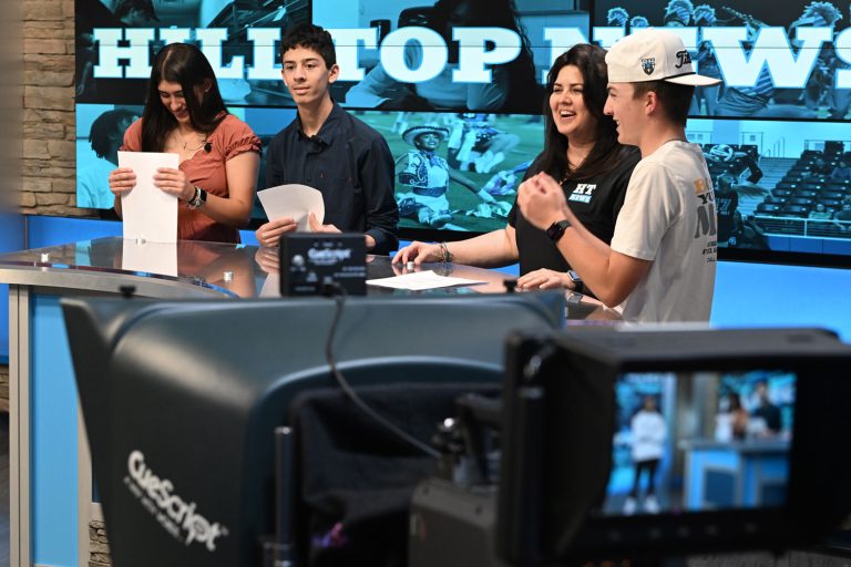 4 staff members of the Hill Top News stand around a news desk laughing candidly.