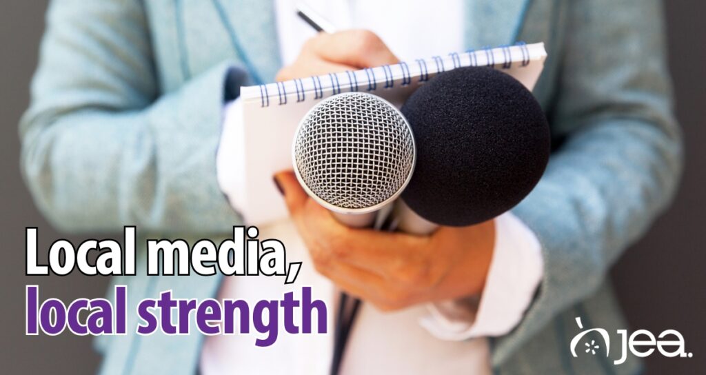 A journalist writing in a notepad holds two microphones. The text reads "Local media, local strength".