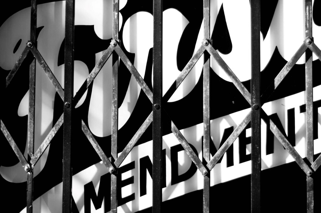 Black and white image containing a tight shot of a fence or gate in front of a sign with the word "First" in white scripted font above the word "AMENDMENT" in all caps and black font with a white fill.