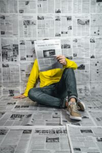 Graphic contains a person wearing a yellow shirt and jeans sitting on the floor, leaning against a wall and reading a newspaper. The floor and wall are covered in newspapers. There is no text in the image.