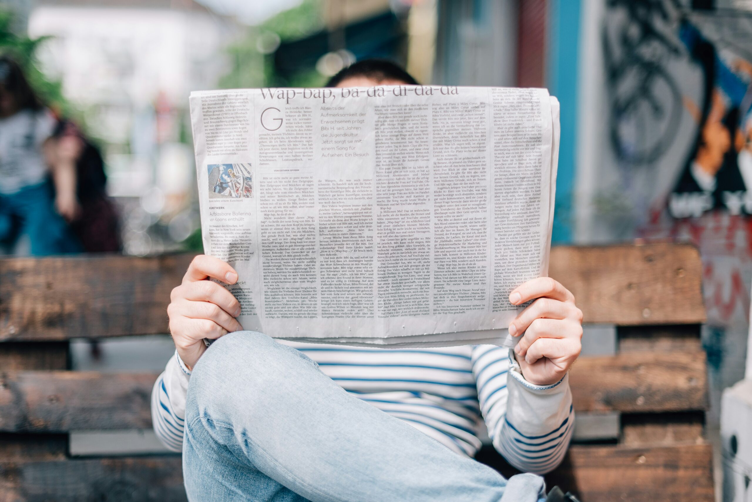 Graphic contains a person wearing a striped shirt and jeans sitting on a bench with crossed legs reading a newspaper. There is no text in the image.
