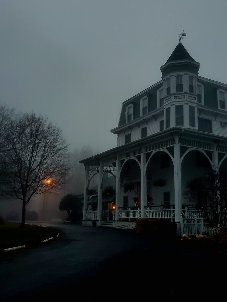 Image contains a spooky old white house with a bare tree to the left and a yellow street light glowing in background.