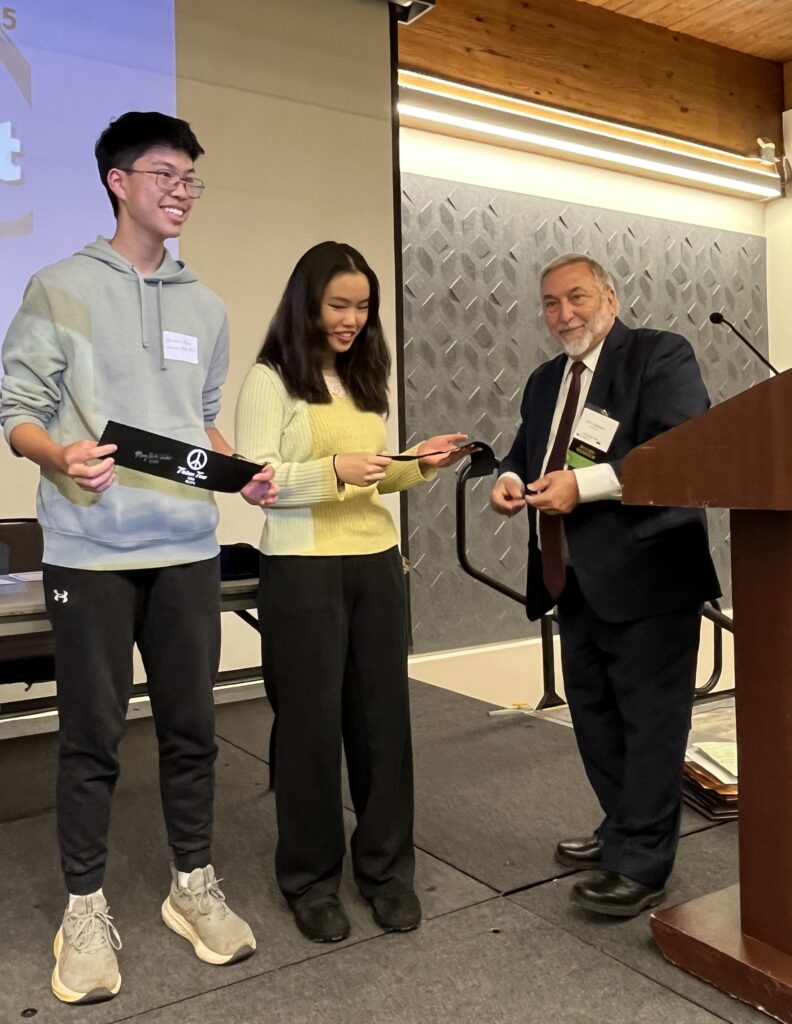 The image shows John Tagliareni in a black suit standing in front of a podium to the right as he presents black armbands to two students, a female (center) and male (left).