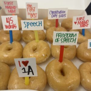 Image contains rows of glazed donuts in a box, each with a different color toothpick with a First Amendment word or phrase handwritten in marker.
