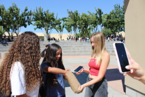 Photo contains a student journalist, right, wearing a red top holding a brown paper bag as a high school student, center, in a blue top draws something from the bag and a third student looks on while a fourth student records on a phone.