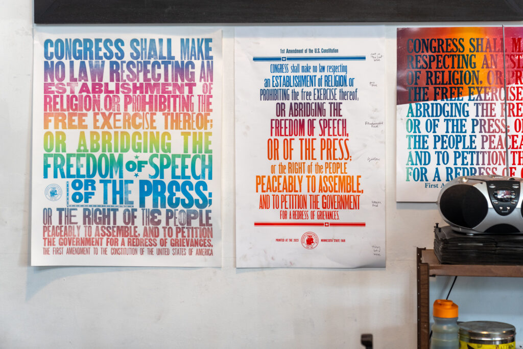 The image is comprised of three posters on a white wall, each containing the 45 words of the First Amendment in bright rainbow colored font. There is a brown wooden shelf with a CD player and water bottle in the bottom right corner of the frame.