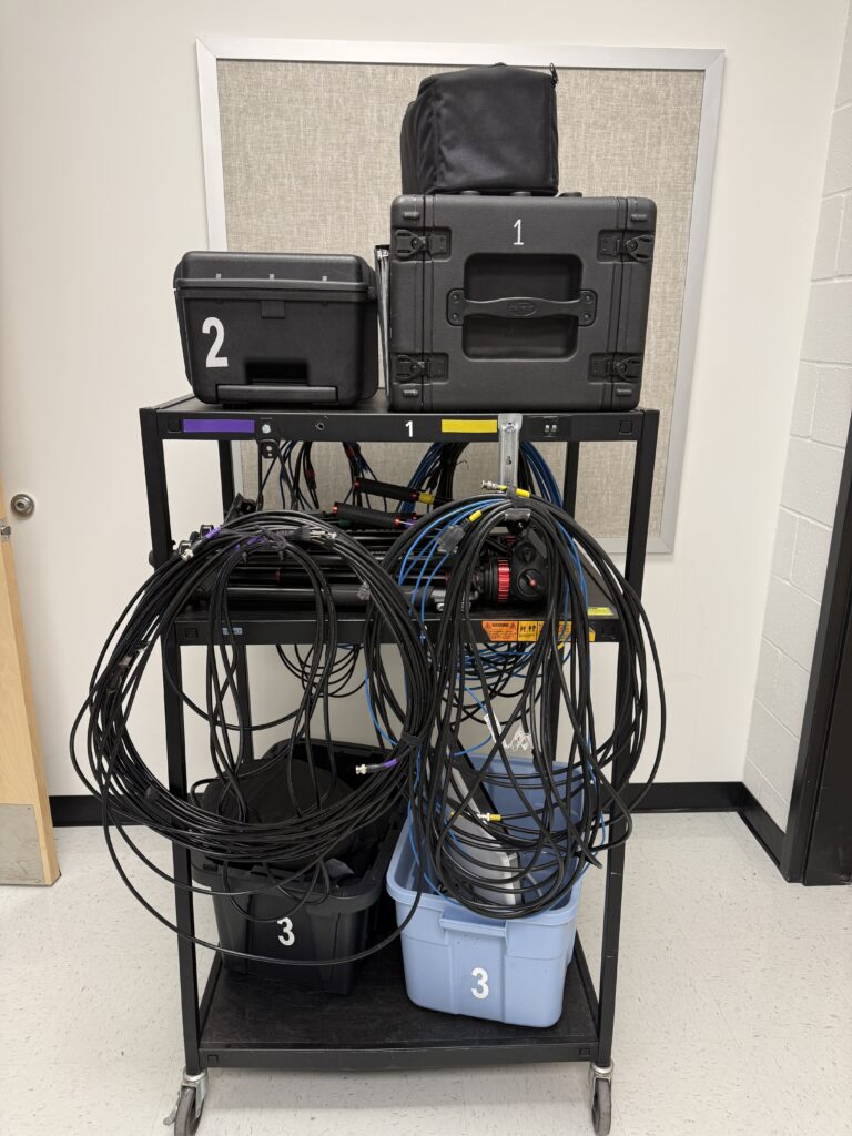 This is a cart with wheels. It has three shelves on it that has boxes and cables organized on it.