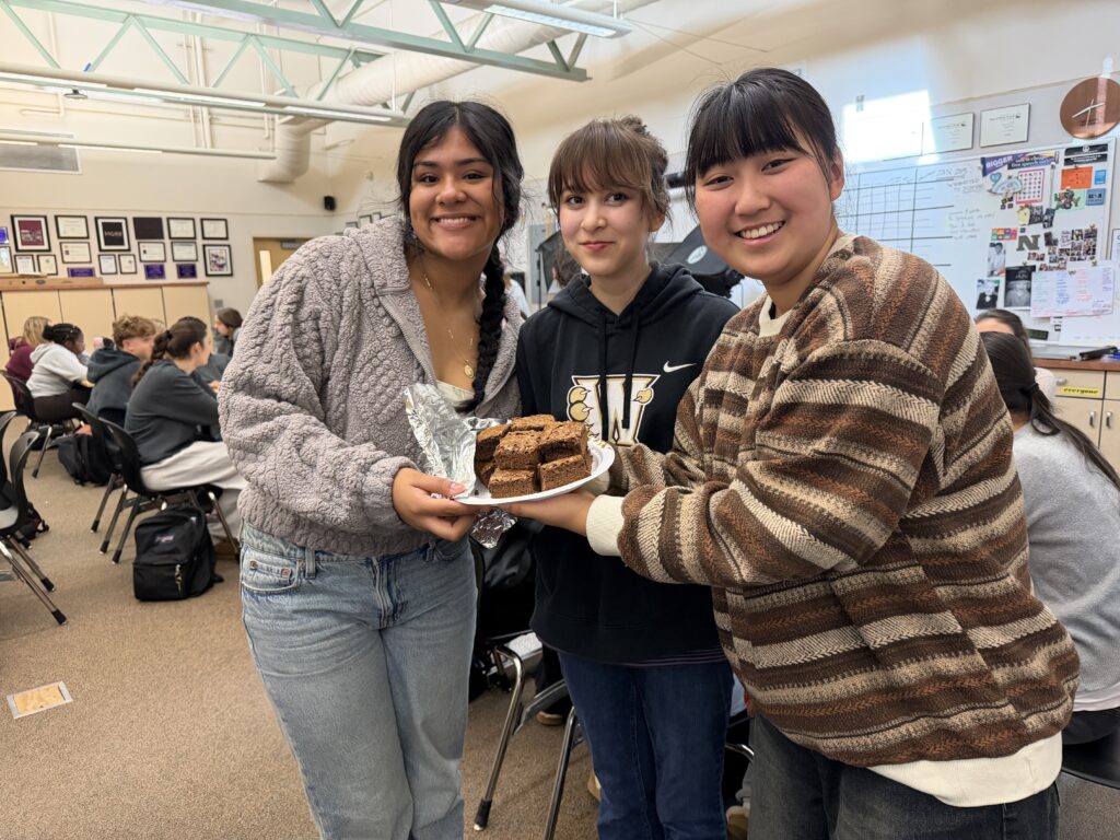 Image depicts three female students holding a paper plate of brownies, as students work in the background.