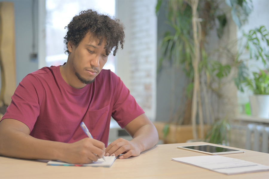 This is an image of a young male in a maroon tshirt writing a letter at a table.