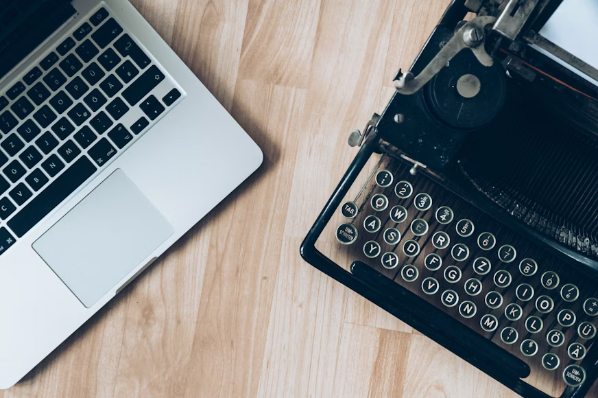 Image shows part of a silver laptop with black keyboard in the upper left corner and a black typewriter in the right corner, both on a light wooden surface.