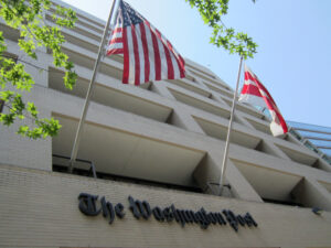 Image shows the front of the Washington Post building with two flags hanging of its sign. 