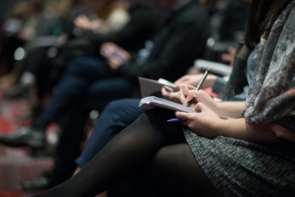 Image shows the laps of several people seated in a row and holding notebooks and taking notes.