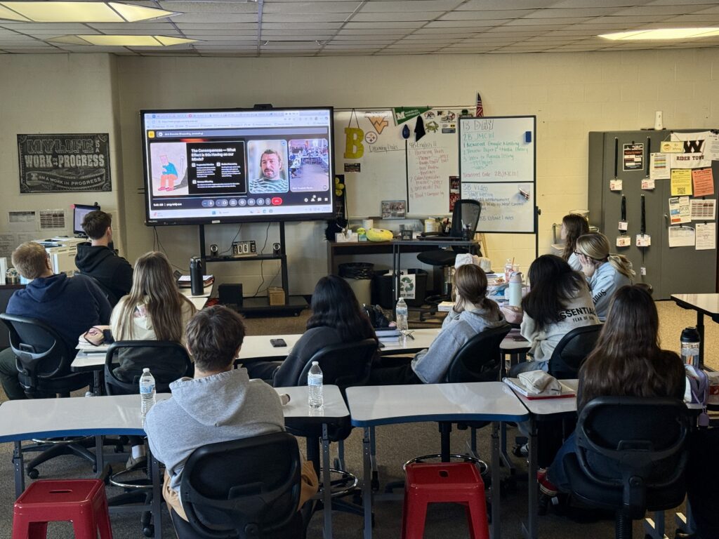 Image depicts students sitting in a classroom looking at a digital display screen with a man's face and text on the screen. The walls contain classroom posters and decor.
