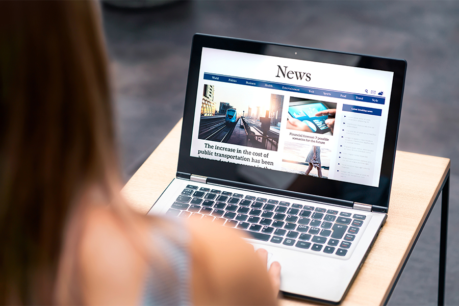 This is an over the shoulder image of a girl looking at a computer screen. On the screen is the word "News" and a news looking website. The laptop is sitting on a small square table, like a school desk.