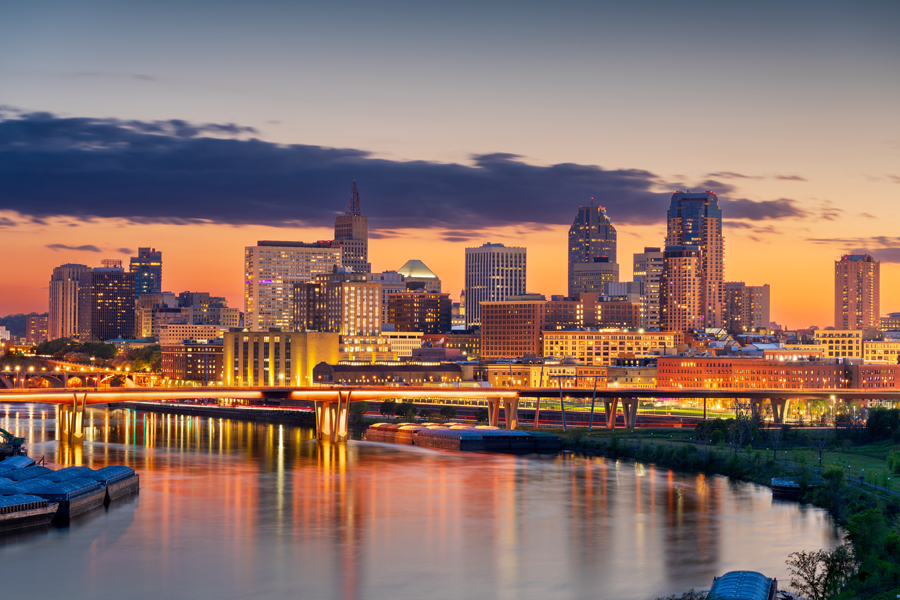 This is an image of the St. Paul/Minneapolis downtown skyline. There is a river in the foreground of the image and tell buildings set in front of a sunset.