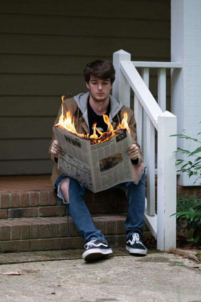 Image depicts a teen boy wearing blue jeans and a hooded sweatshirt sitting on red brick steps, leaning on a white railing, and reading a newspaper that is on fire.