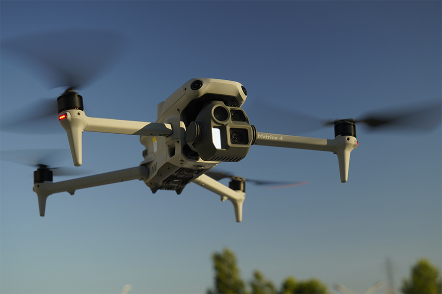 A drone helicopter hovers above the ground in front of a blue sky.