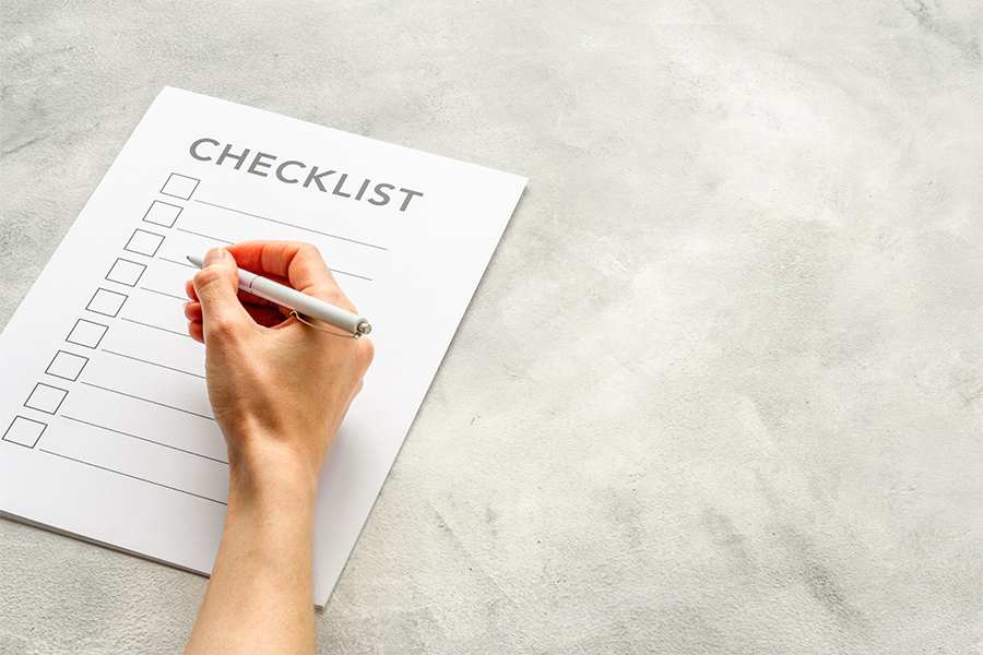 Decorative image of a piece of paper on a table with the word checklist on it that has a hand on top of the list preparing to write something.